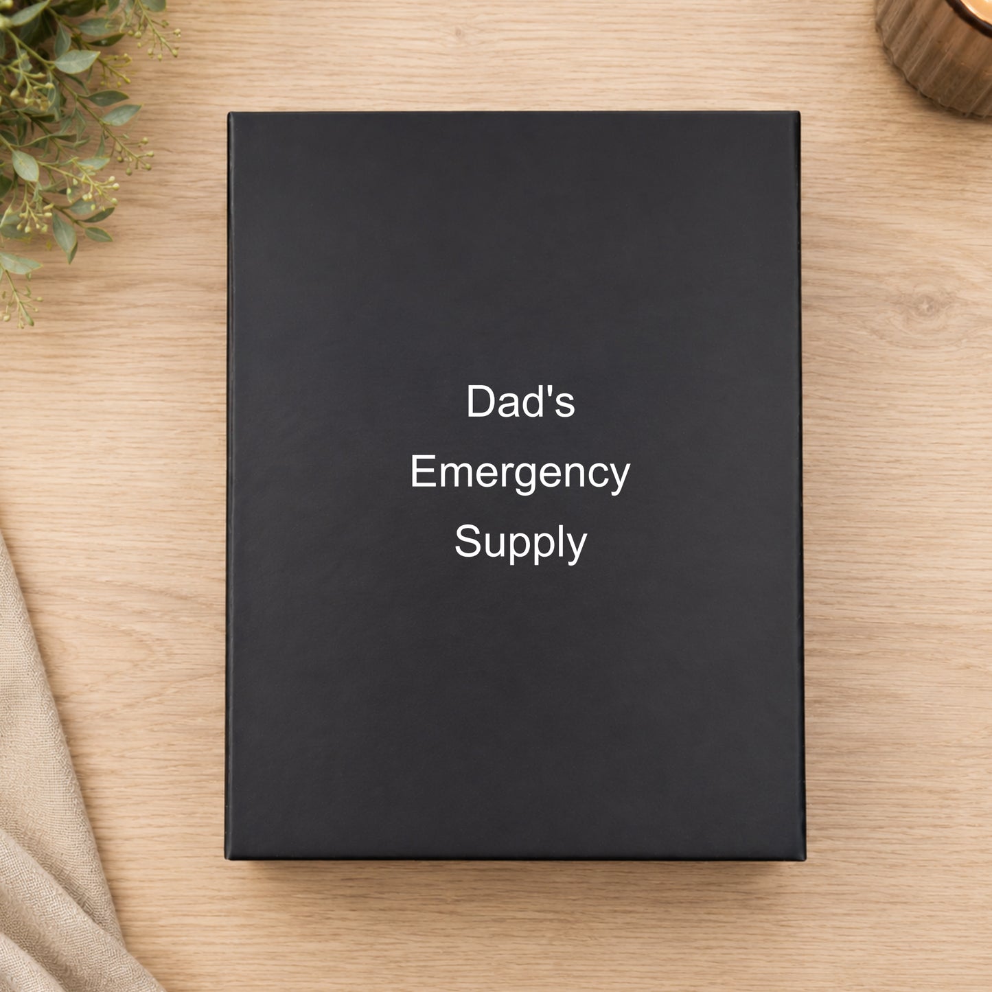 Black box labeled 'Dad's Emergency Supply' on a wooden surface with a plant and fabric.