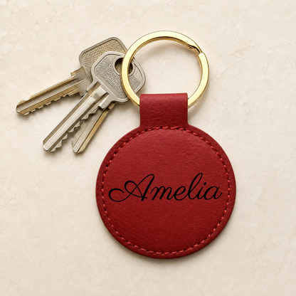 A personalized round red leather keychain with the name 'Amelia' embroidered in black script, attached to a gold split ring with two keys, on a light beige textured background.