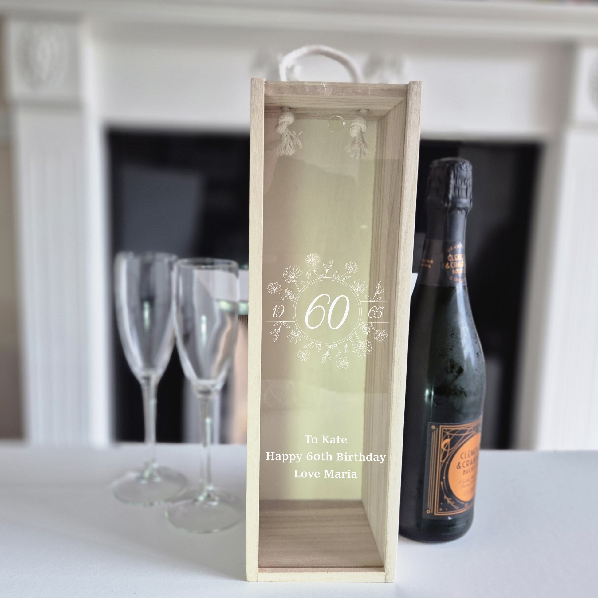60th birthday champagne bottle box next to a bottle and 2 glass. The cover to the box is clear acrylic with white writing on saying 60 and a message underneath