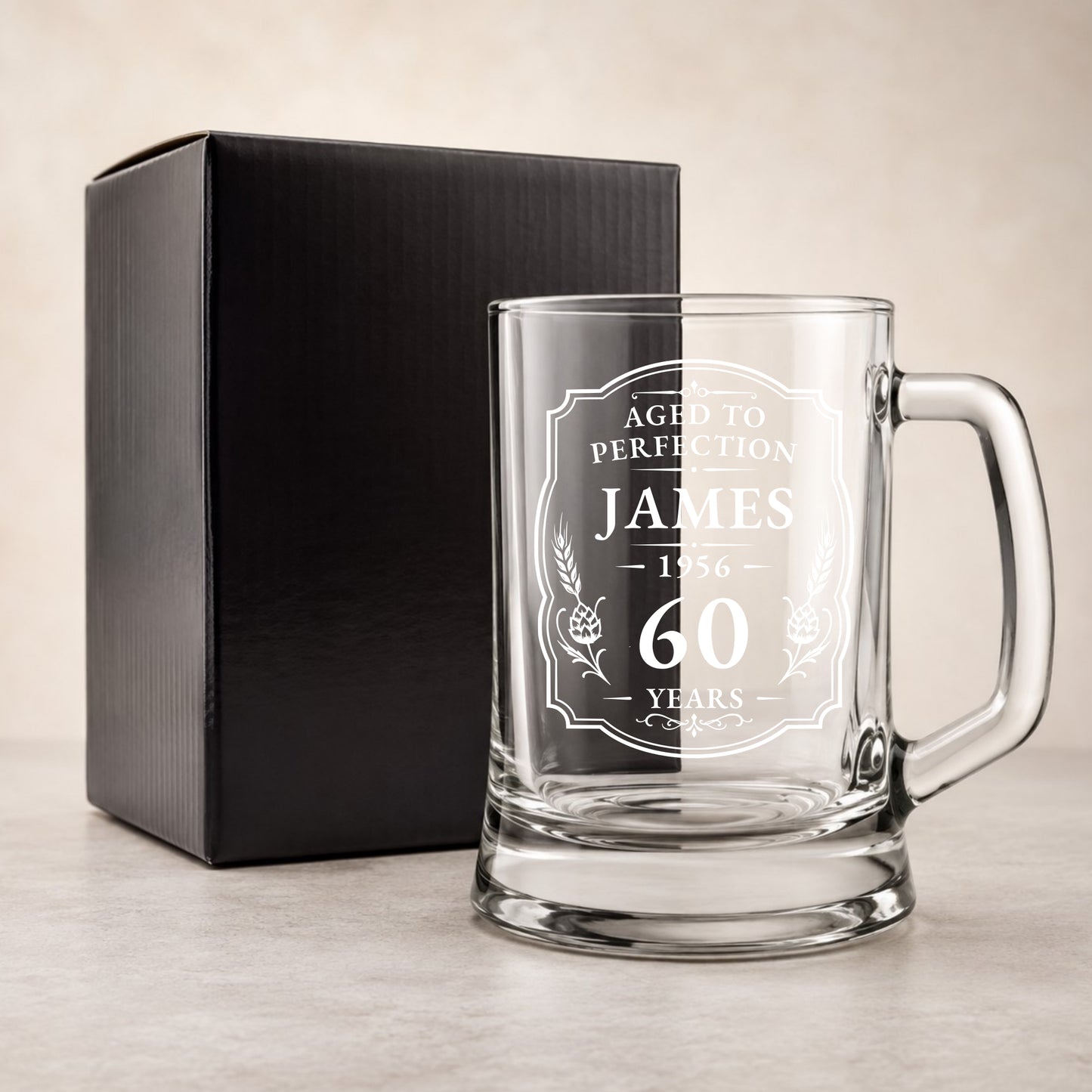 Clear glass mug with engraved text and a black gift box on a beige background