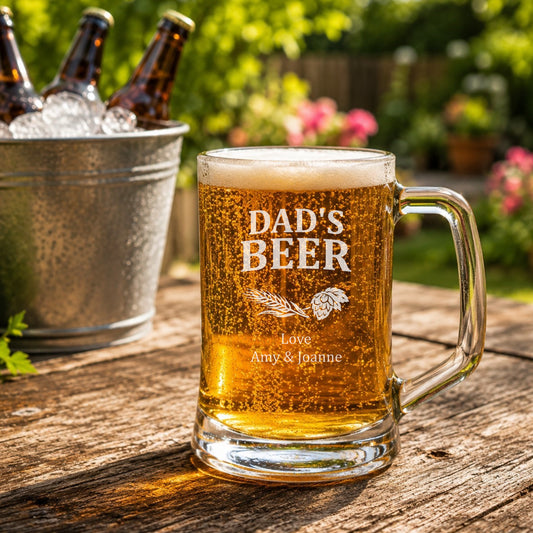 Glass mug of beer with 'DAD'S BEER' engraving on a wooden table outdoors.