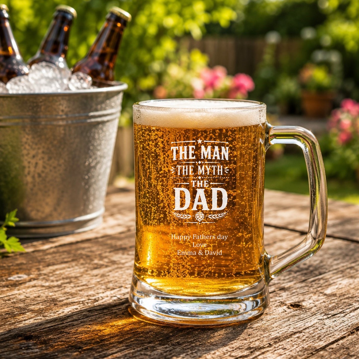 Glass mug of beer with 'The Man, The Myth, The Dad' text on a wooden table outdoors.
