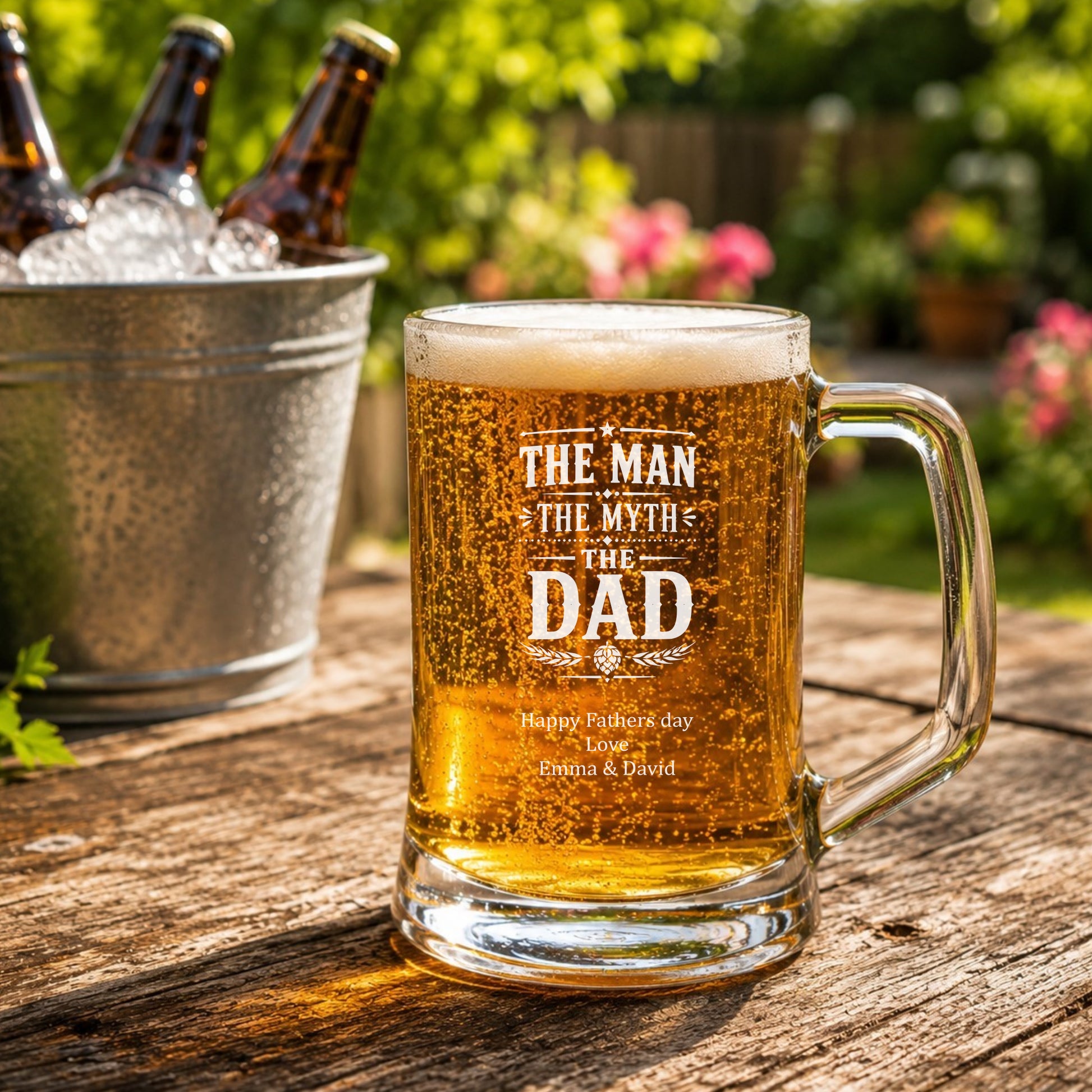 Glass mug of beer with 'The Man, The Myth, The Dad' text on a wooden table outdoors.