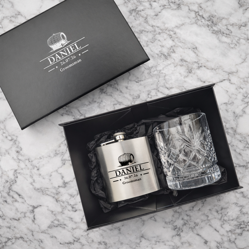 Personalized hip flask and whisky glass set in a black gift box on a marble surface.