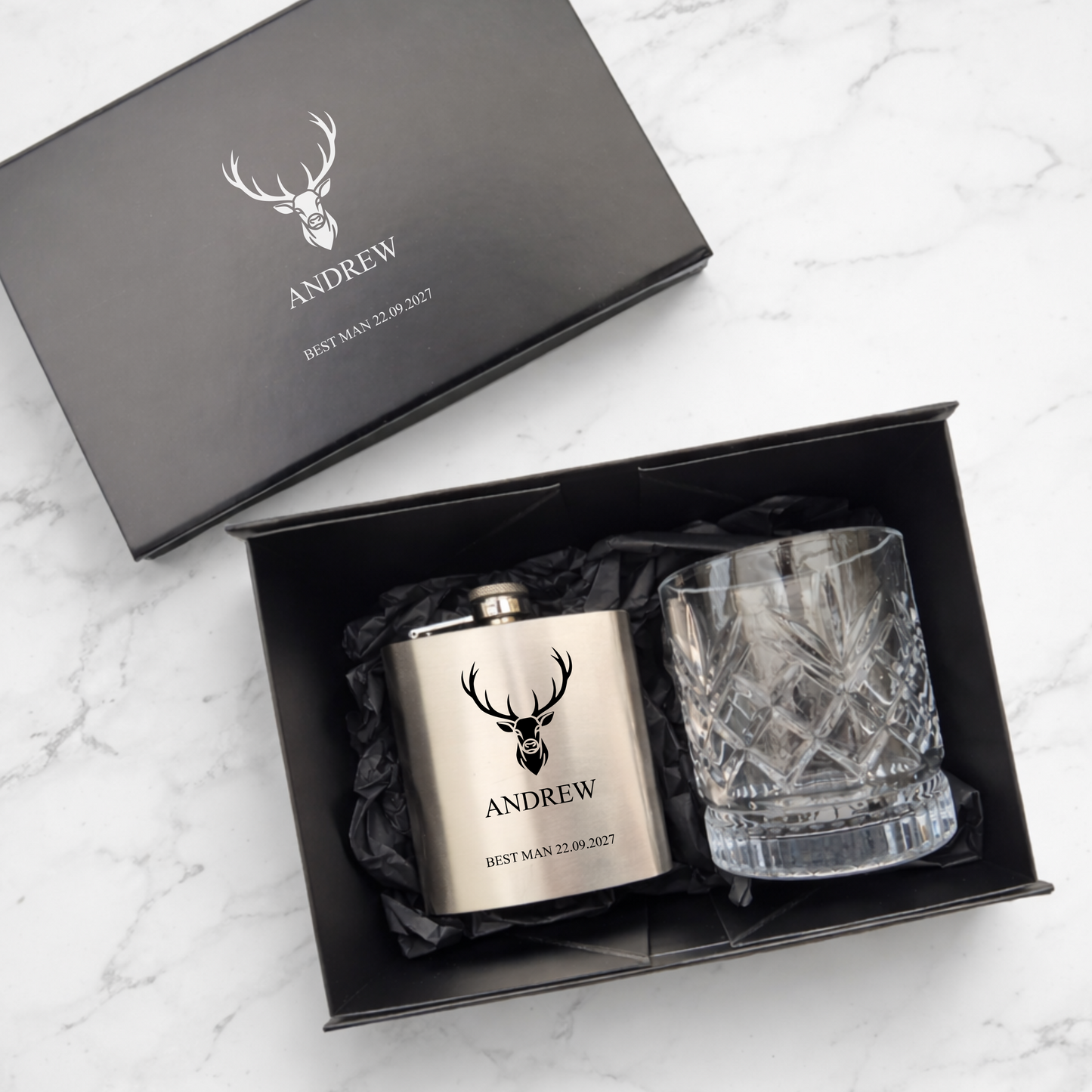 Personalized hip flask and glass set with 'ANDREW' engraving in a black gift box on a marble surface.