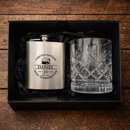 Engraved hip flask and crystal glass set in a black box on a wooden surface.