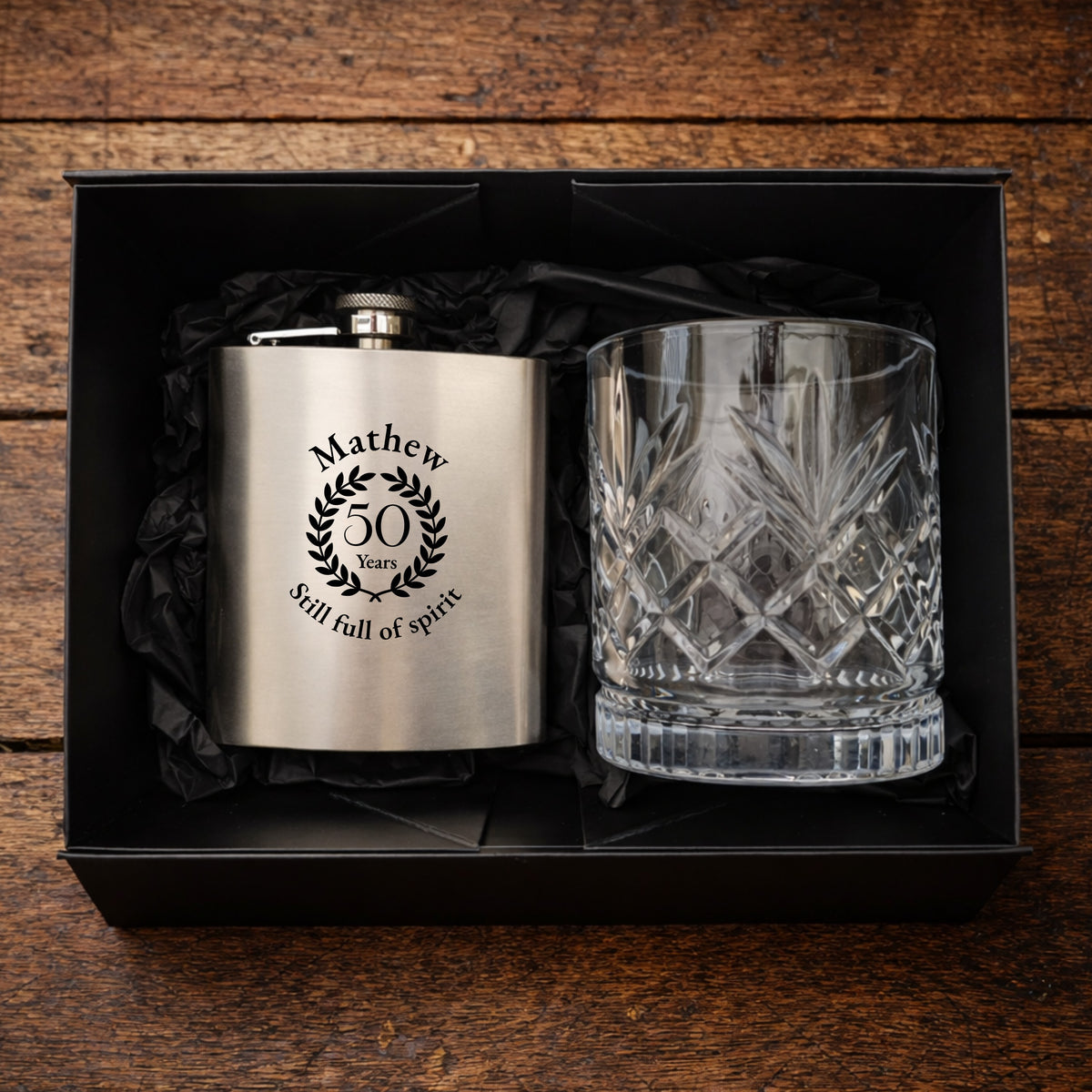 Engraved hip flask and crystal glass set in a black box on a wooden surface.