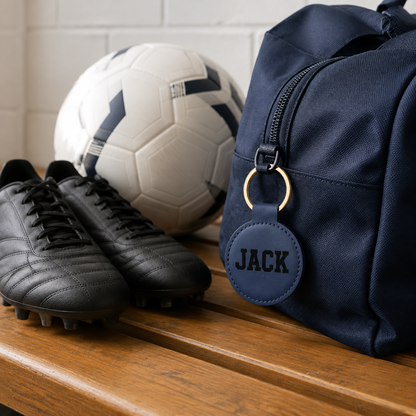 A navy blue sports duffel bag with a personalized round leather keychain engraved with the name 'JACK', styled on a wooden bench next to black soccer cleats and a white soccer ball.