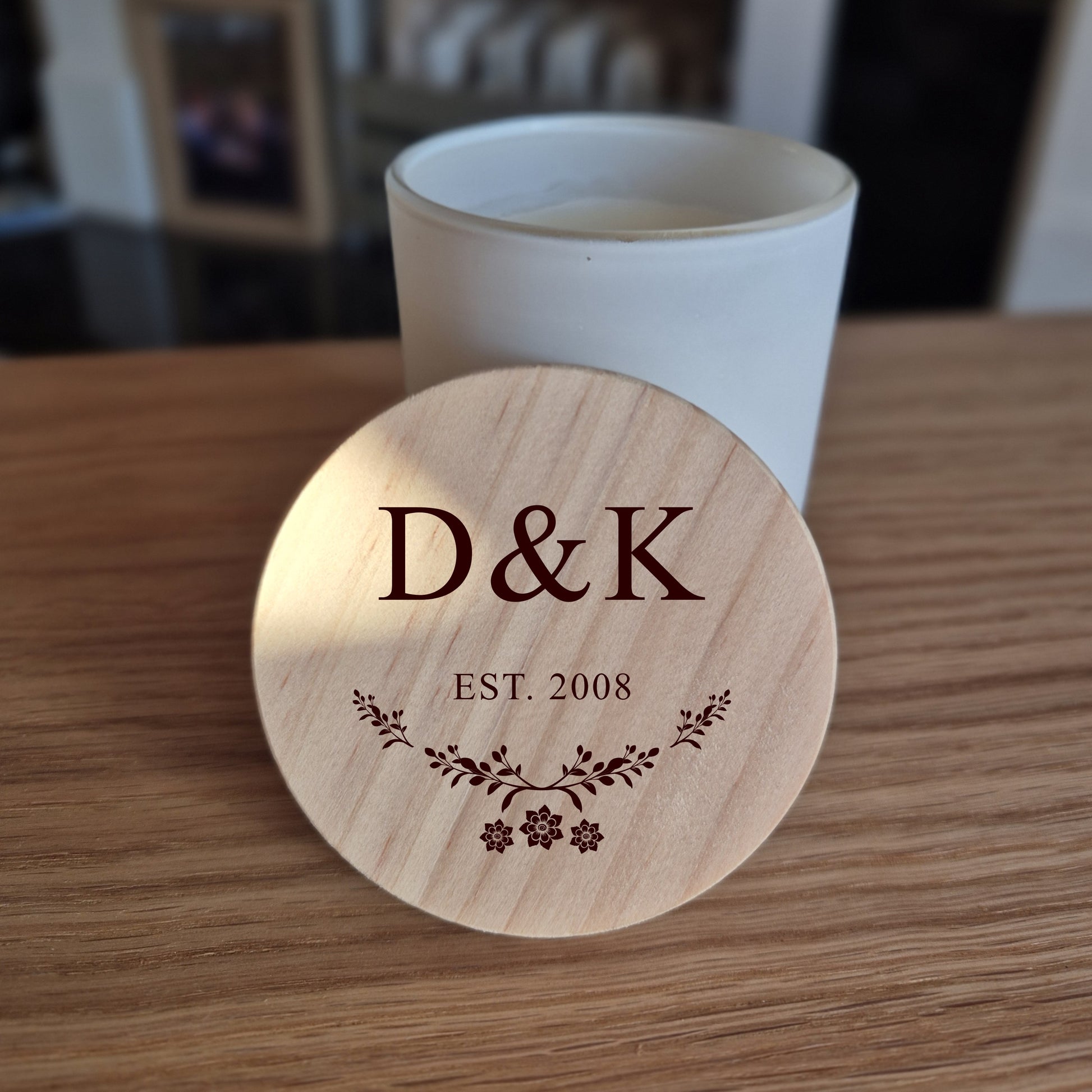 White jar candle with engraved wooden lid featuring 'D&K EST. 2008' on a wooden surface.