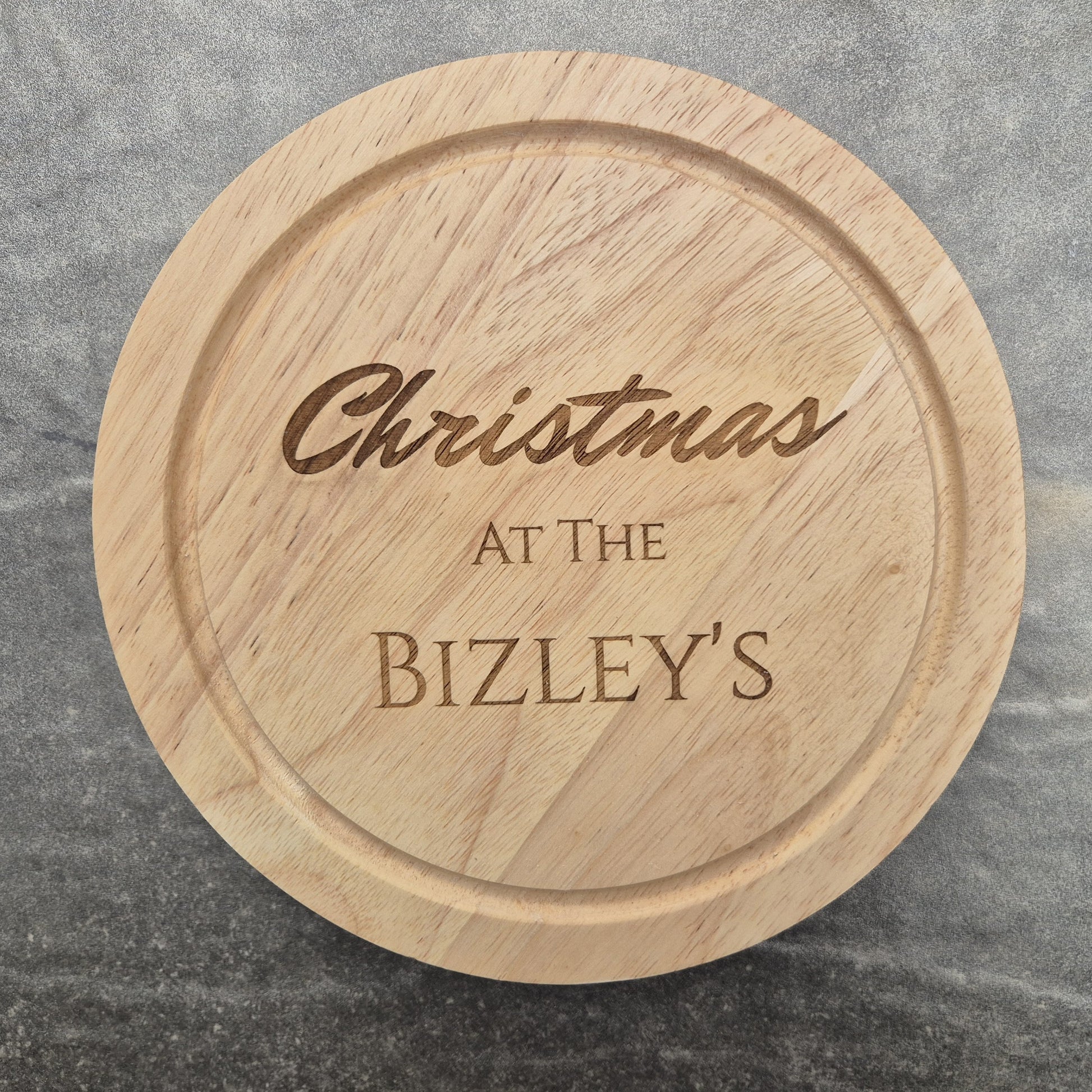Personalised cheese board with engraved text 'Christmas at The Bizley's' on a gray surface