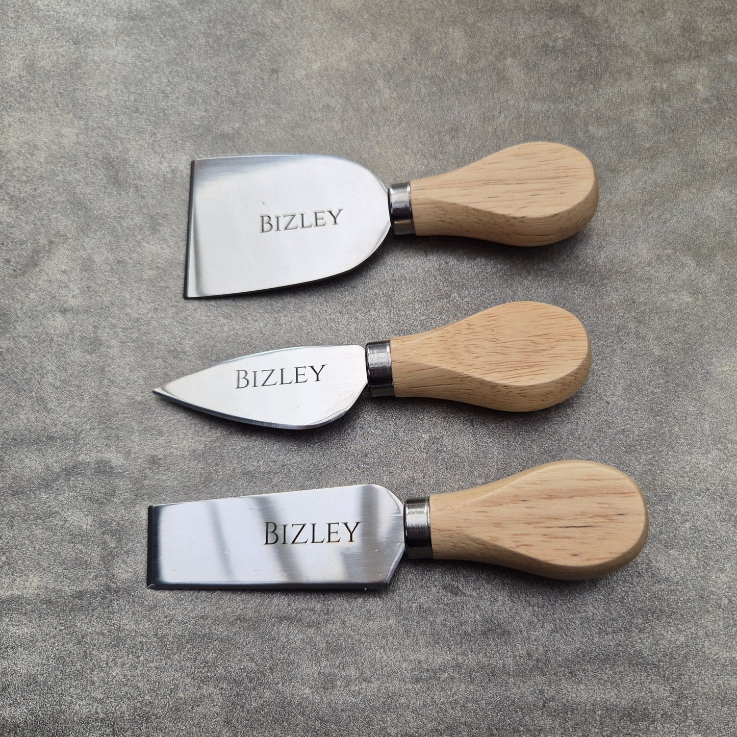 Three cheese knives with wooden handles and metal blades on a gray surface, branded 'BIZLEY'.