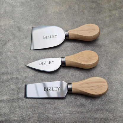 Three cheese knives with wooden handles and metal blades on a gray surface, branded 'BIZLEY'.