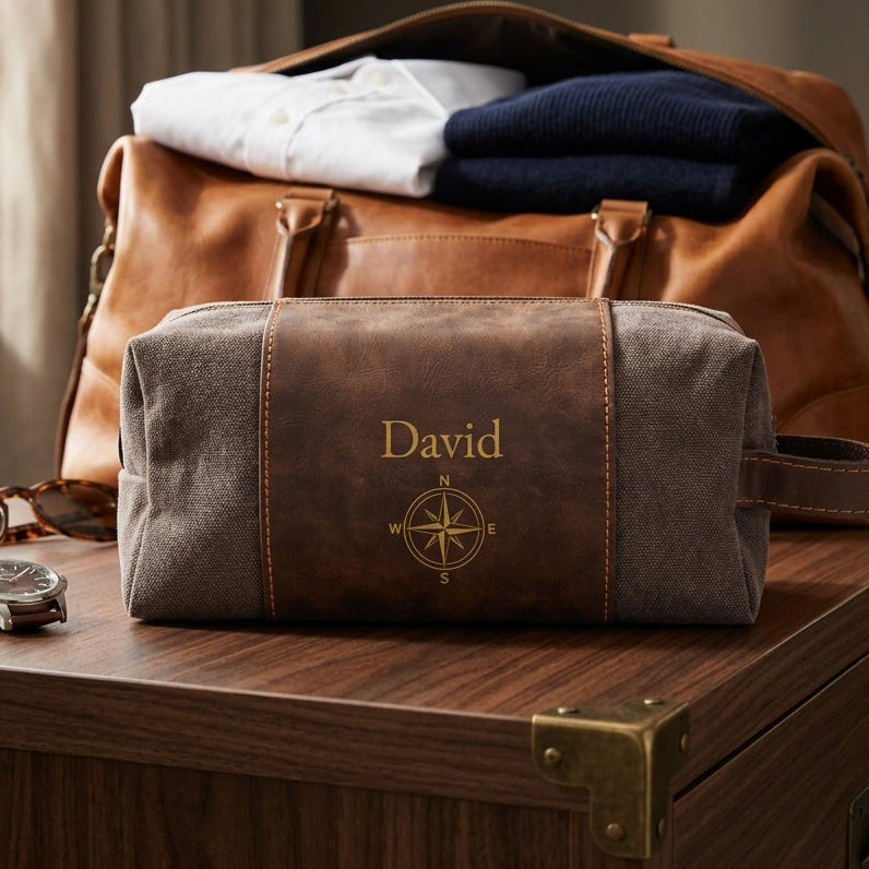 A personalized brown leather and gray canvas travel toiletry bag with the name 'David' and a compass design, displayed on a wooden chest in front of a brown leather duffel bag.