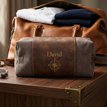 A personalized brown leather and gray canvas travel toiletry bag with the name 'David' and a compass design, displayed on a wooden chest in front of a brown leather duffel bag.