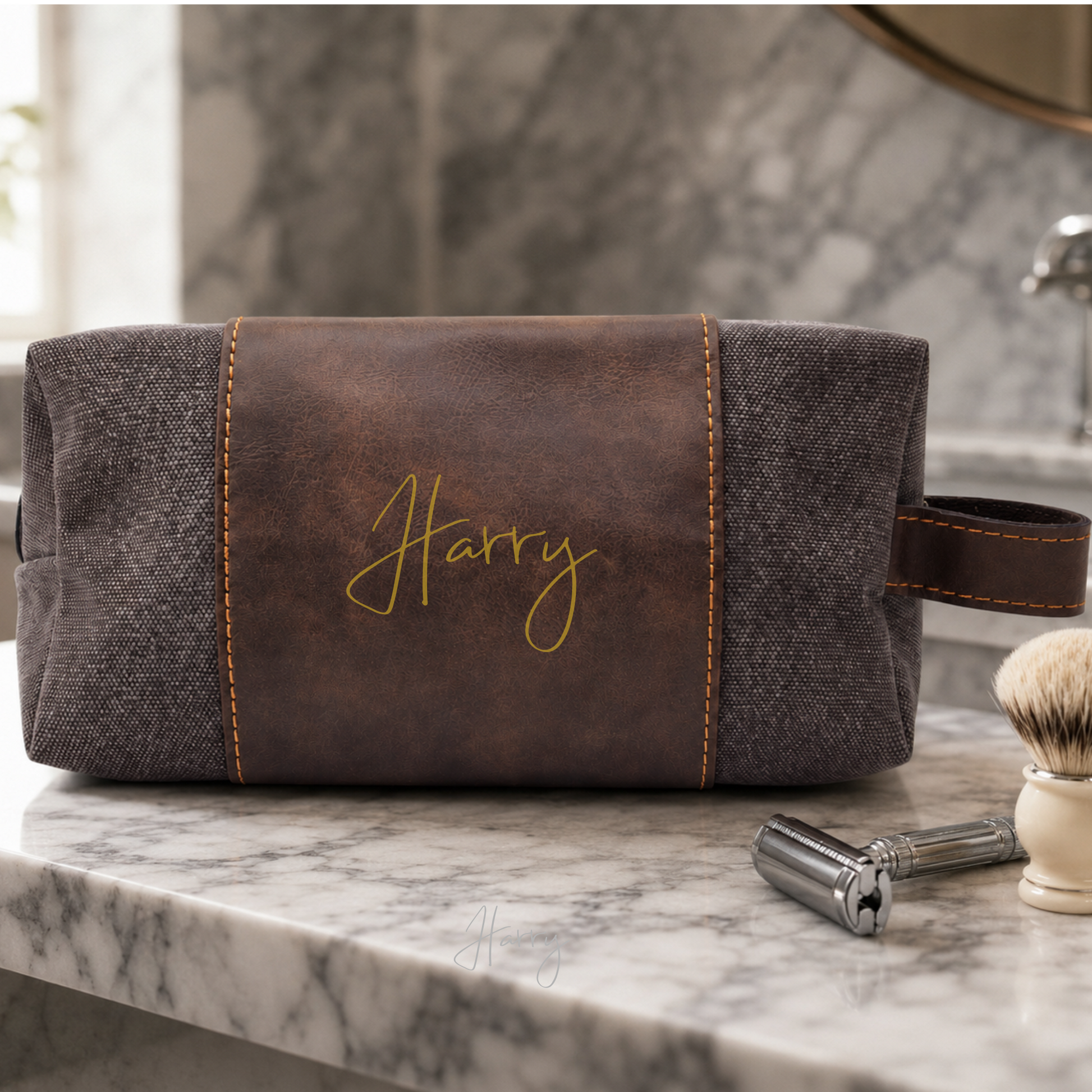 A personalized men's travel grooming pouch with a brown faux-leather center panel and dark gray textured fabric sides, featuring the name 'Harry' embroidered in gold on the front. The pouch is styled on a marble countertop with a shaving brush and razor nearby.