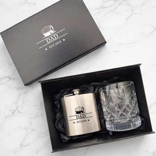 Gift set with hip flask and whiskey glass in a black box labeled 'DAD EST. 2010' on a white marble background.