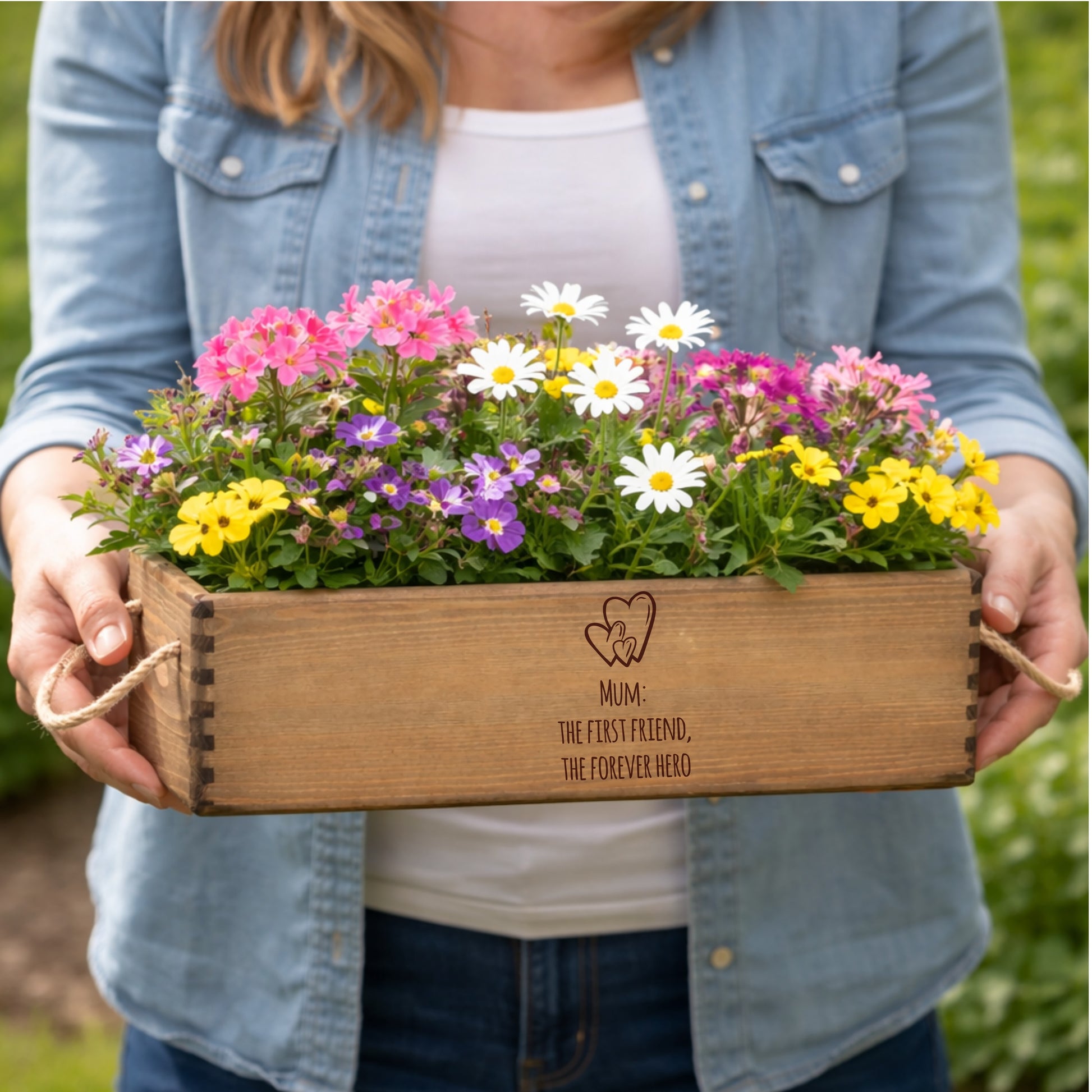 Person holding a wooden planter box with flowers and 'Mum: The first friend, the forever hero' engraving.