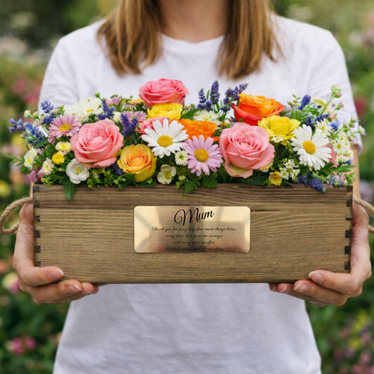 Person holding a wooden box of flowers with 'Mum' engraved, outdoors.