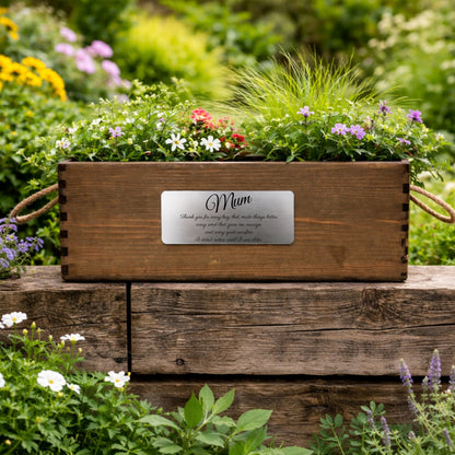 Wooden planter box with flowers and a 'Mum' plaque on a wooden surface with a garden background.