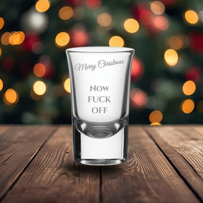 Clear shot glass with printed text on a wooden surface with a blurred Christmas tree background