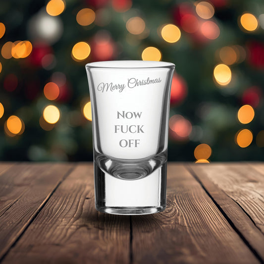 Clear shot glass with printed text on a wooden surface with a blurred Christmas tree background