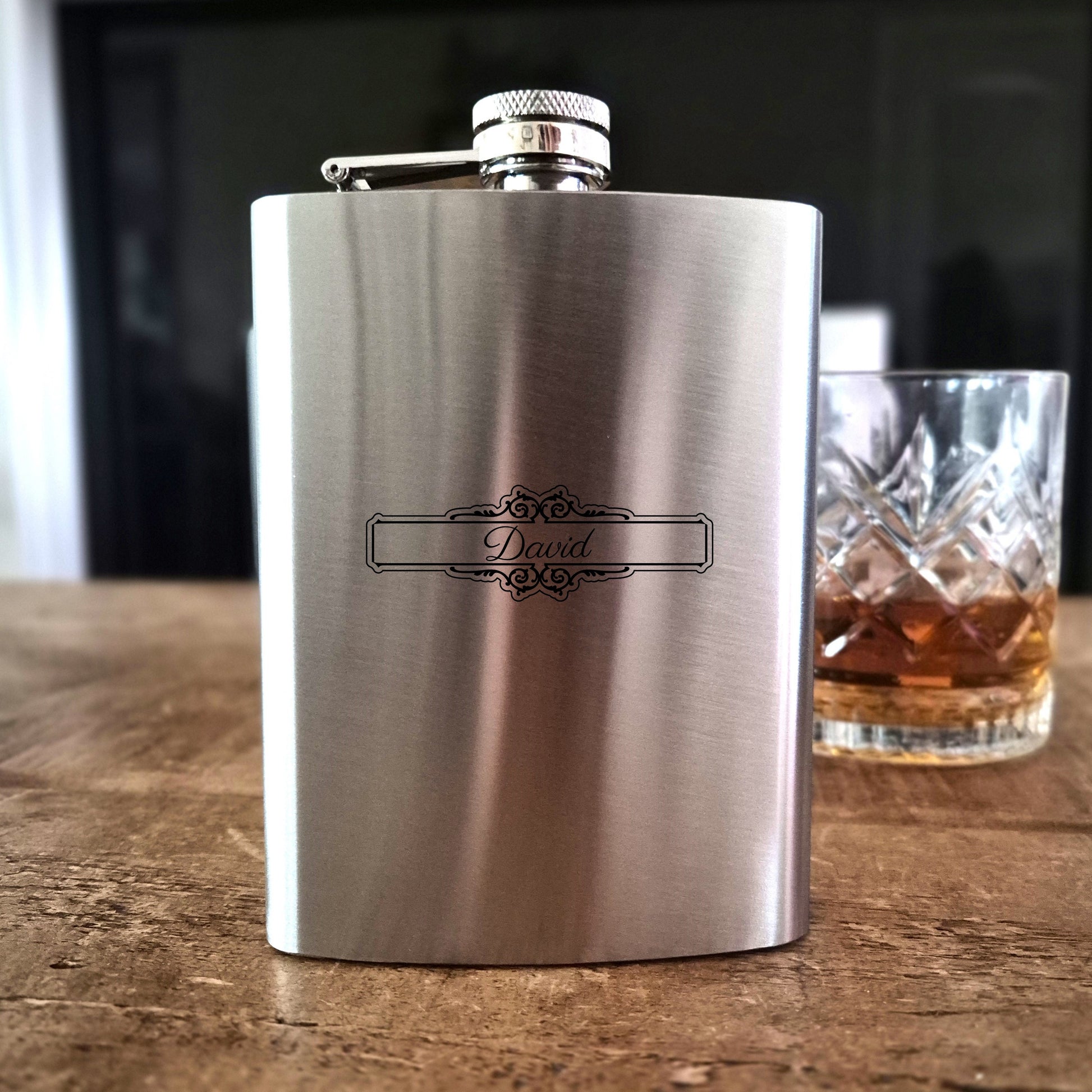 Stainless steel hip flask with 'David' engraving on a wooden surface next to a glass of whiskey.