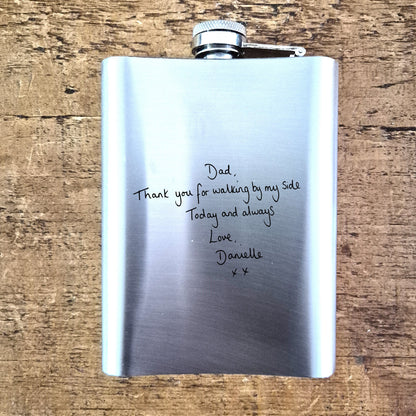 Stainless steel hip flask with handwritten message on a wooden surface