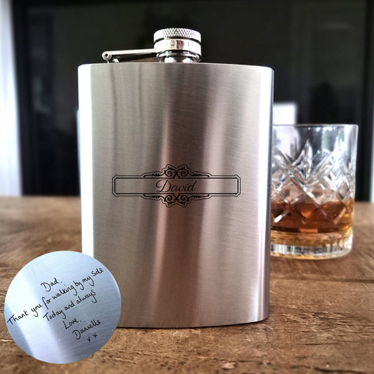Stainless steel flask with engraved name on a wooden surface with a glass of whiskey.