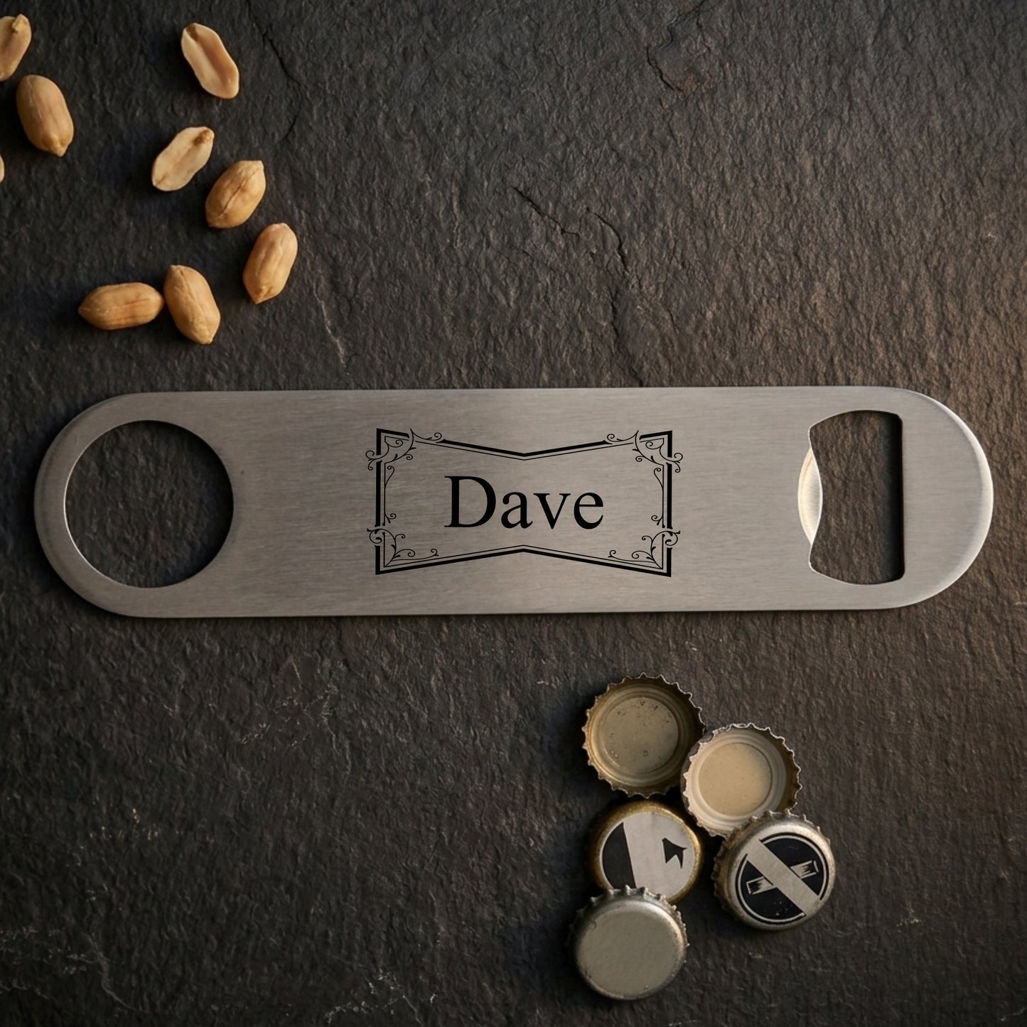 A personalized stainless steel bottle opener engraved with the name 'Dave', displayed on a dark slate background with scattered peanuts and bottle caps.
