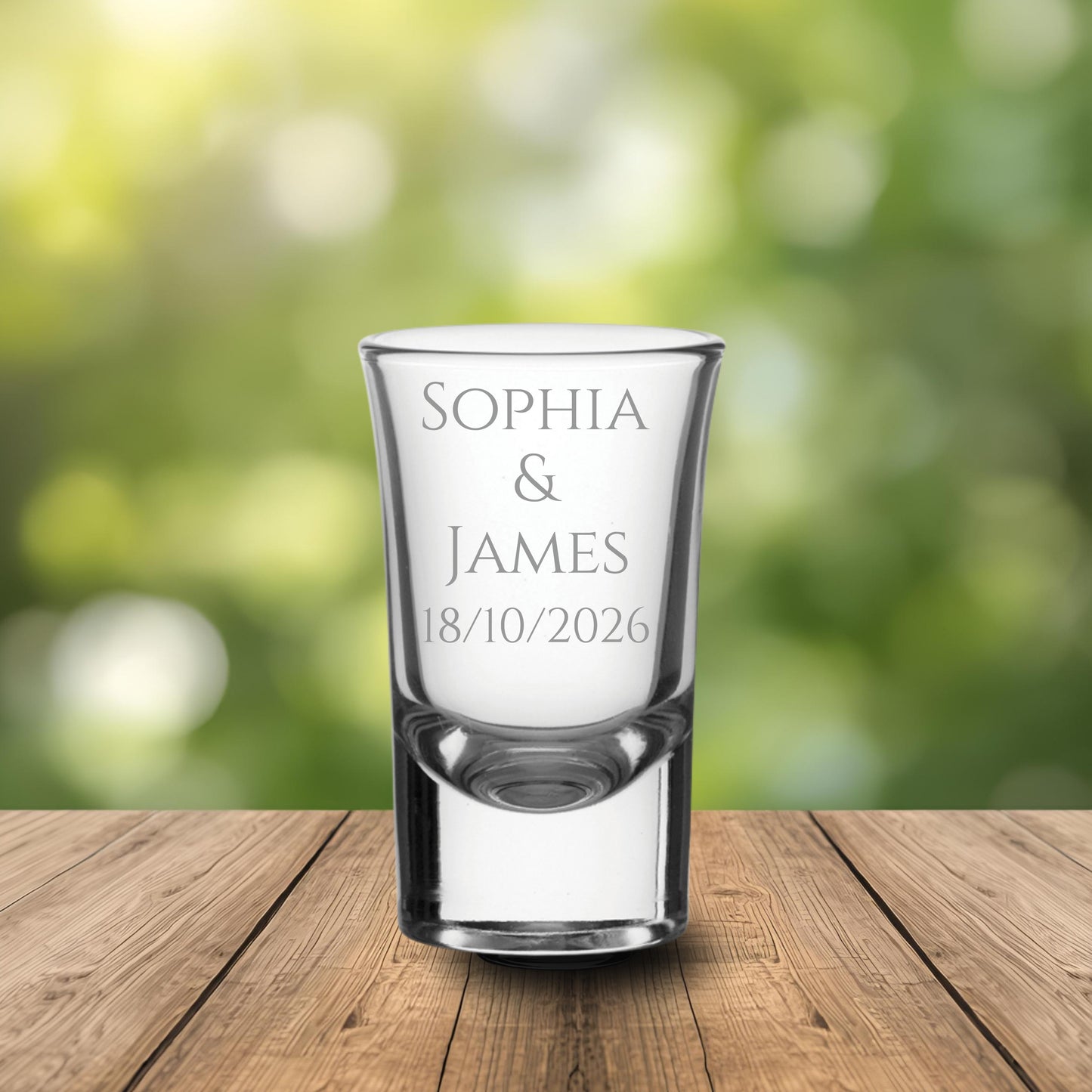 Clear personalised shot glass with engraved text on a wooden surface and blurred green background