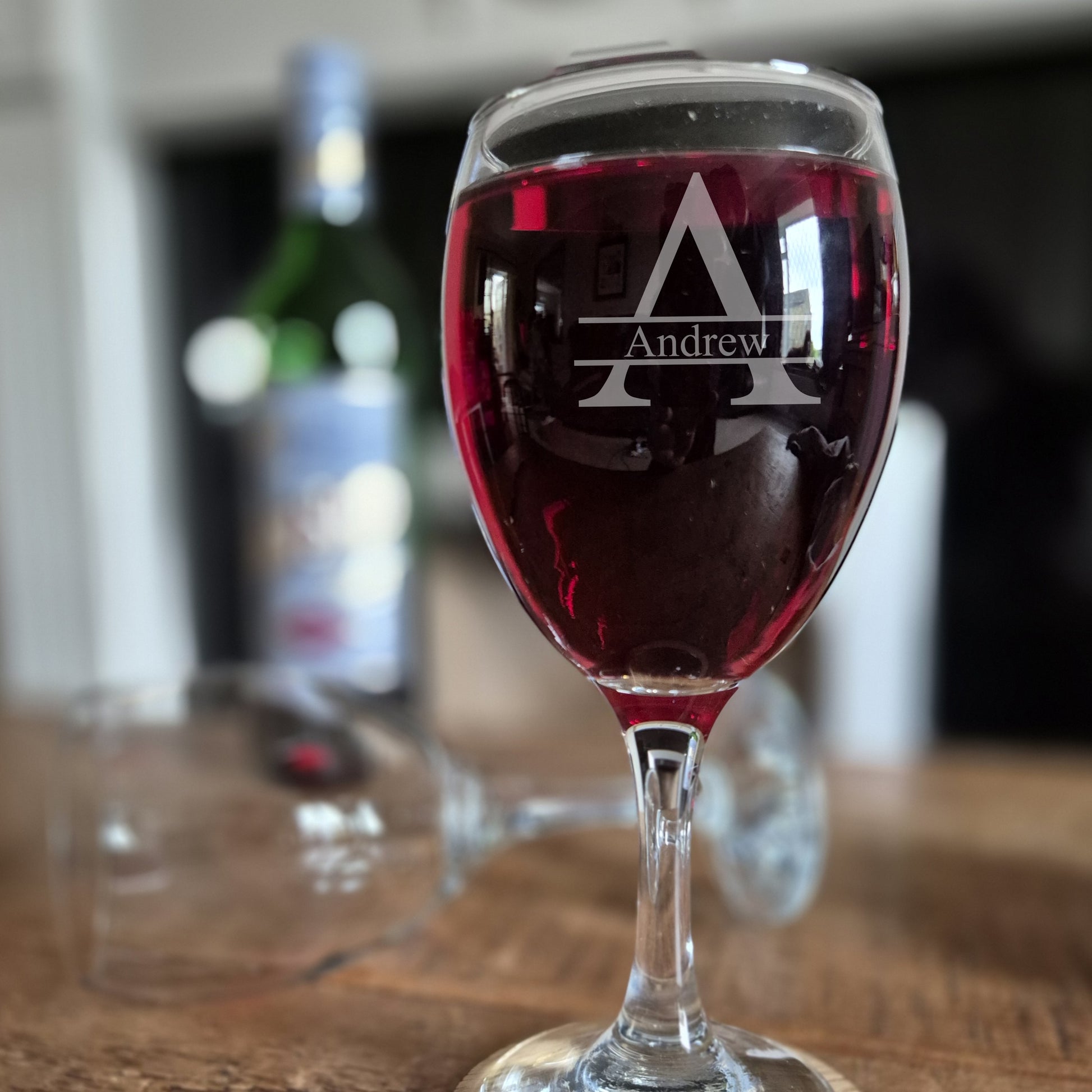 Wine glass with 'Andrew' monogram filled with red wine on a wooden surface.