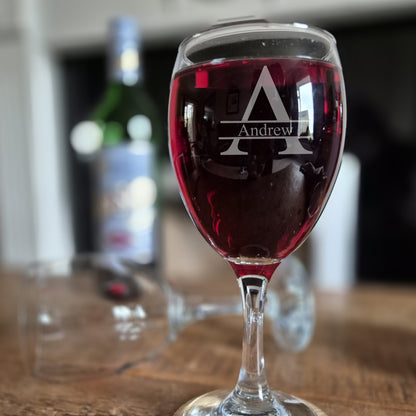 Wine glass with 'Andrew' monogram filled with red wine on a wooden surface.