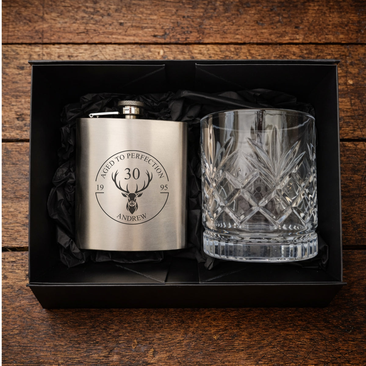 Engraved hip flask and crystal glass set in a black box on a wooden surface.