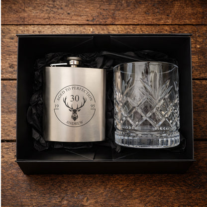Engraved hip flask and crystal glass set in a black box on a wooden surface.