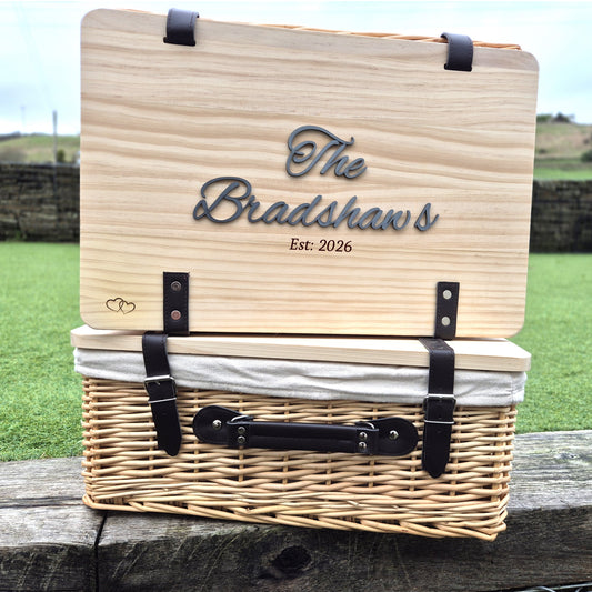 Wicker picnic basket with wooden top engraved with 'The Bradshaws Est. 2026' on a grassy background.