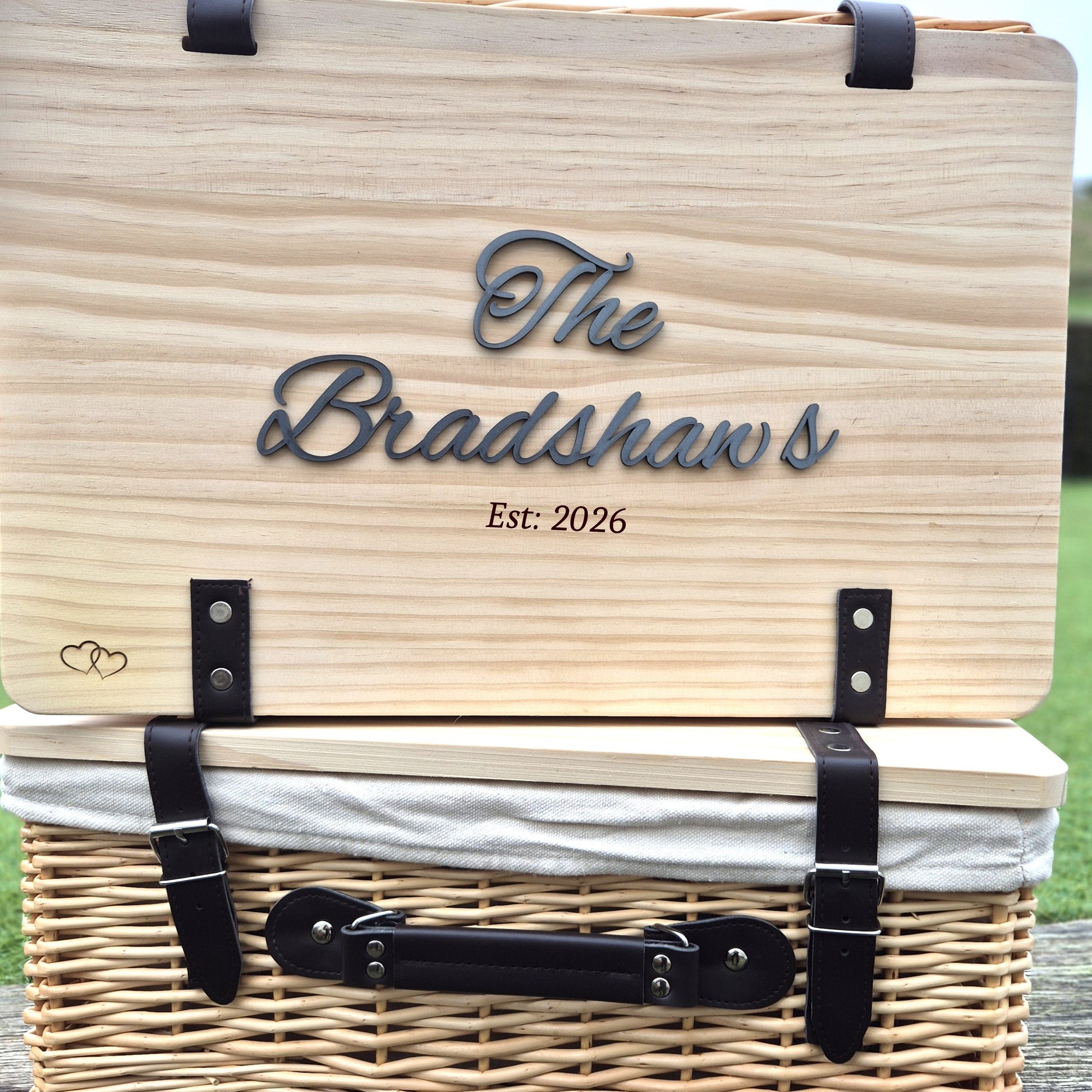 Wooden picnic basket with 'The Bradshaws Est: 2026' engraving on a grassy background