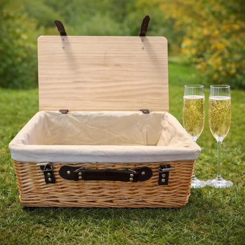 Wicker picnic basket with a white interior on grass, accompanied by two glasses of champagne.