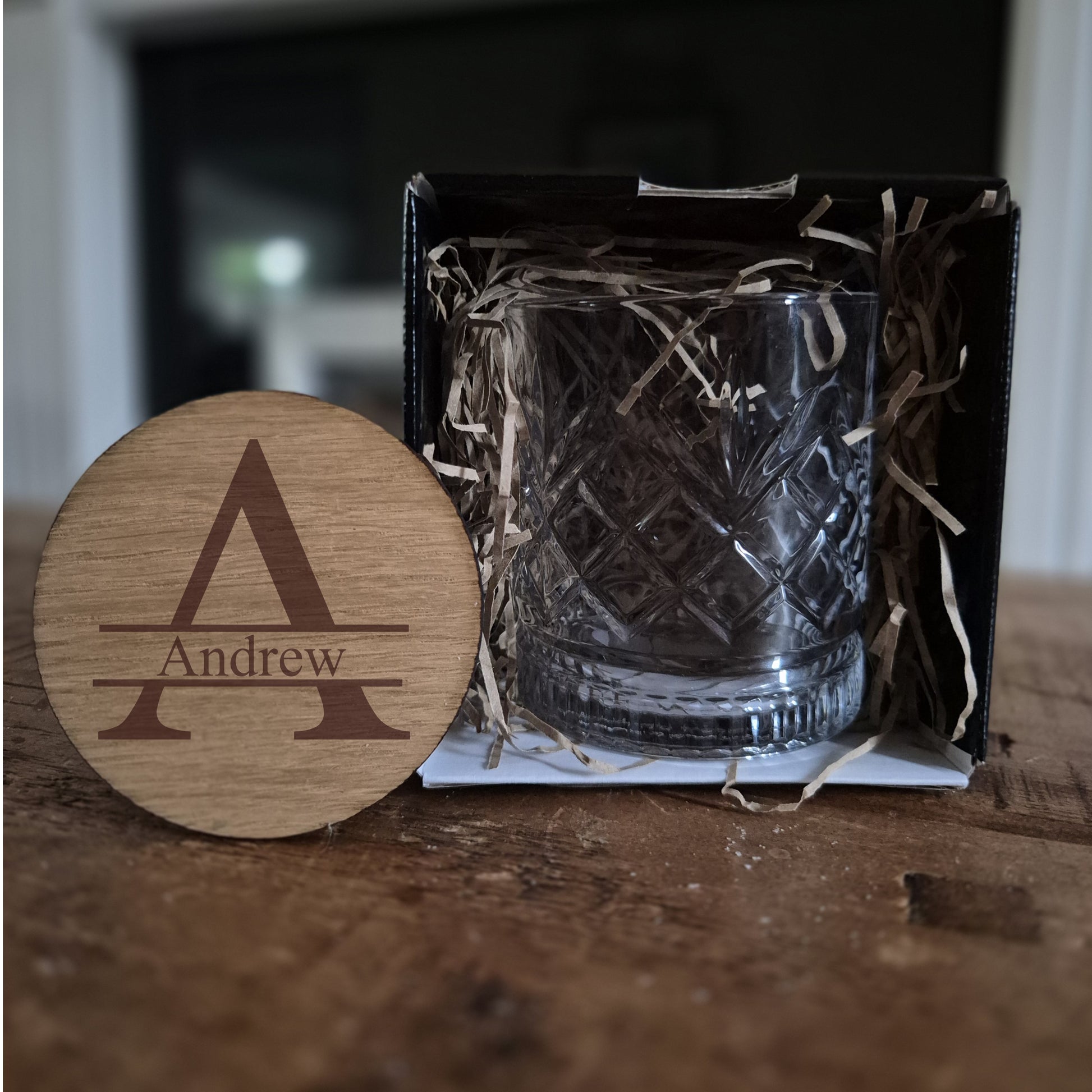 personalised whisky glass in a black box with straw, next to a wooden coaster with 'Andrew' engraved.