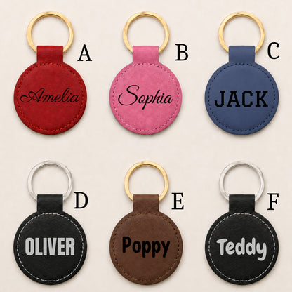 Six personalized round keychains arranged in a 2x3 grid on a light beige background. Each keychain has a stitched leather oval tag with a gold-tone or silver-tone key ring and a custom name or text printed on it.